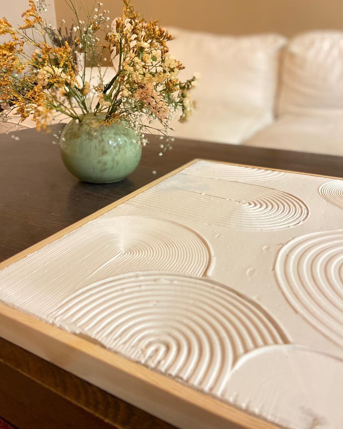 Cuadro blanco con relieve de arcos y círculos sobre una mesa, junto a un jarrón con flores secas