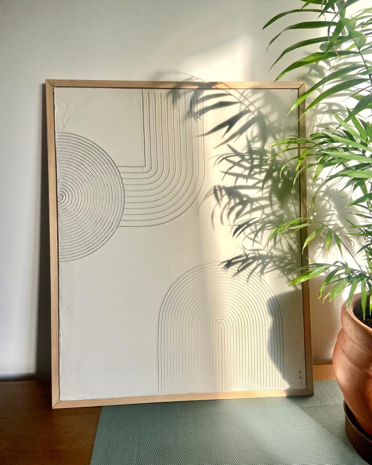 Cuadro blanco con relieve de arcos y círculos, con marco de madera junto a una planta